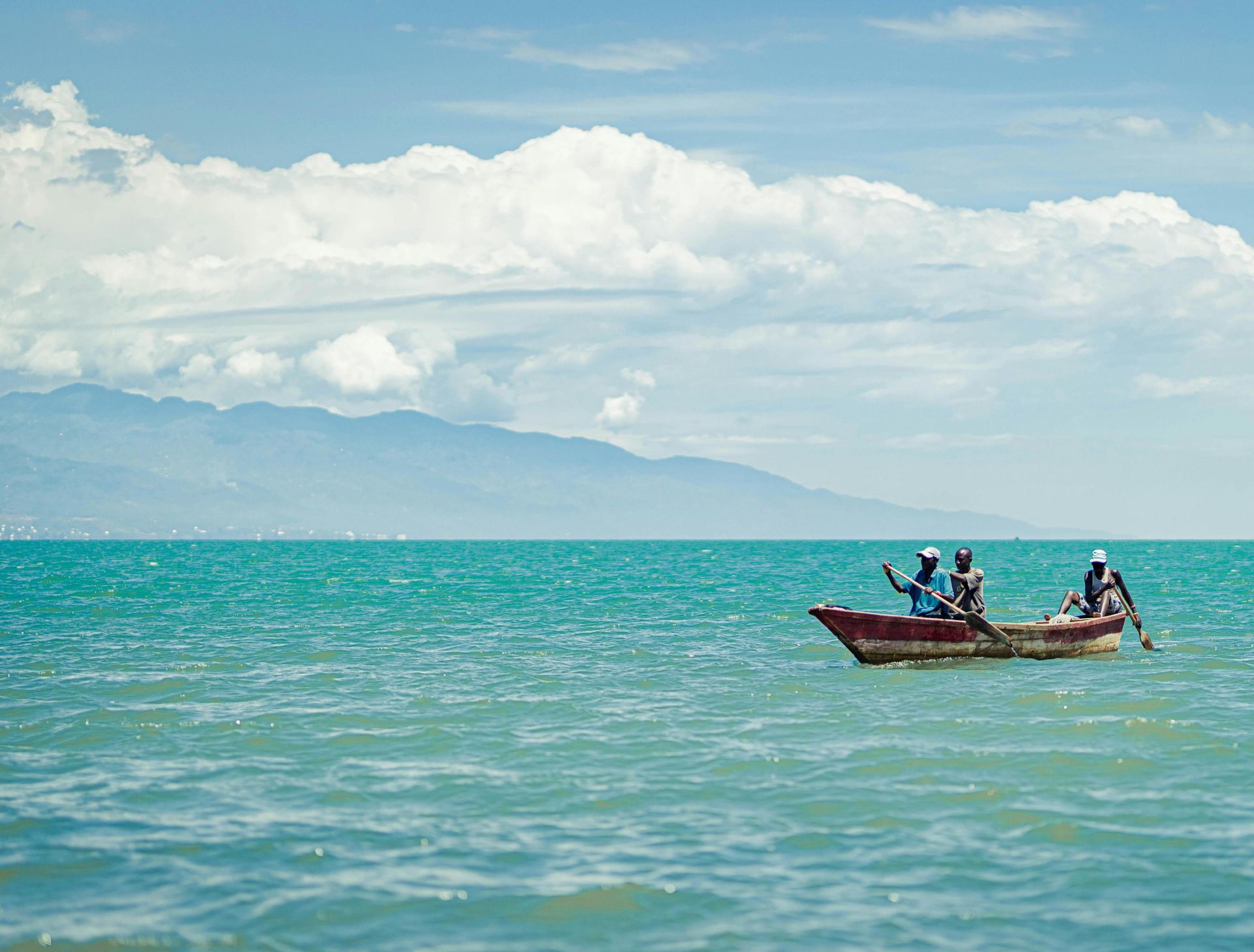 Three individuals in a wooden boat paddling through clear turquoise ocean under a bright sky.